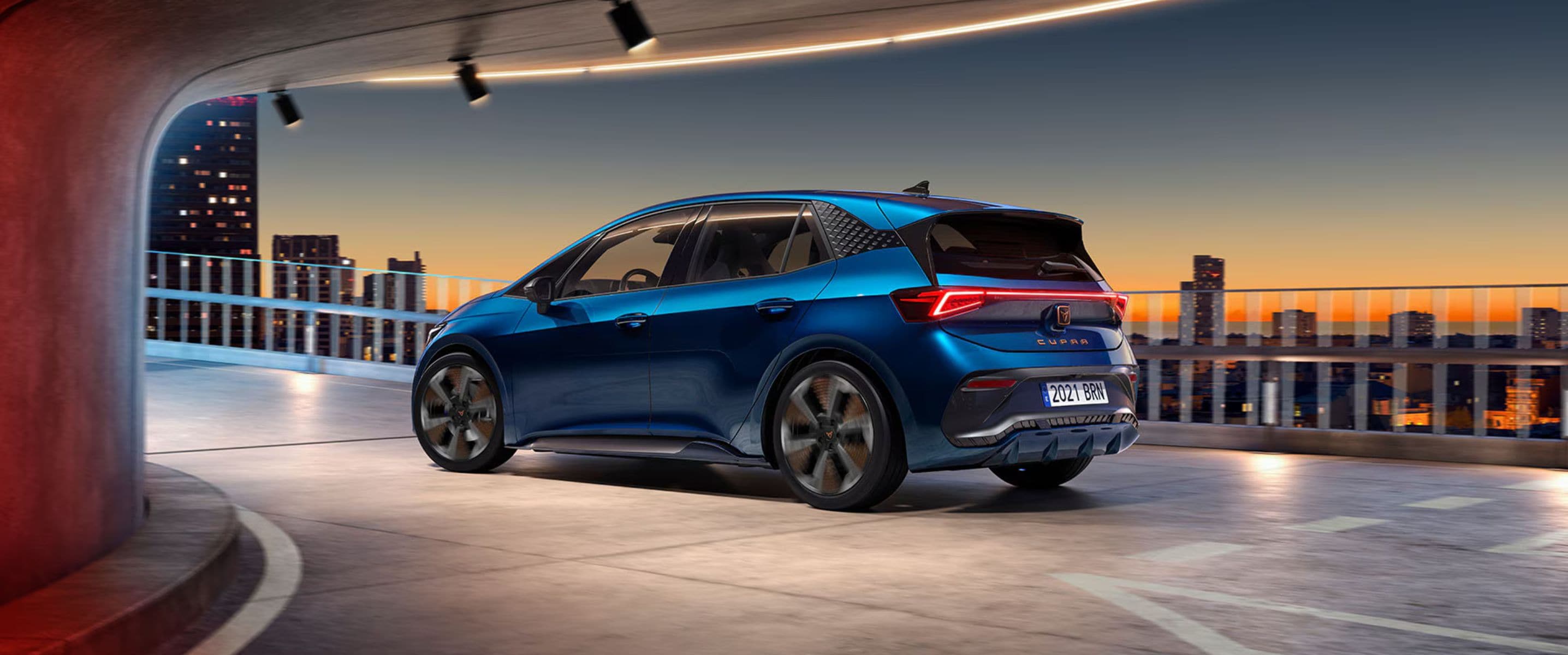 CUPRA Born V3 in Aurora Blue side profile driving up a ramp with Barcelona sunset skyline behind
