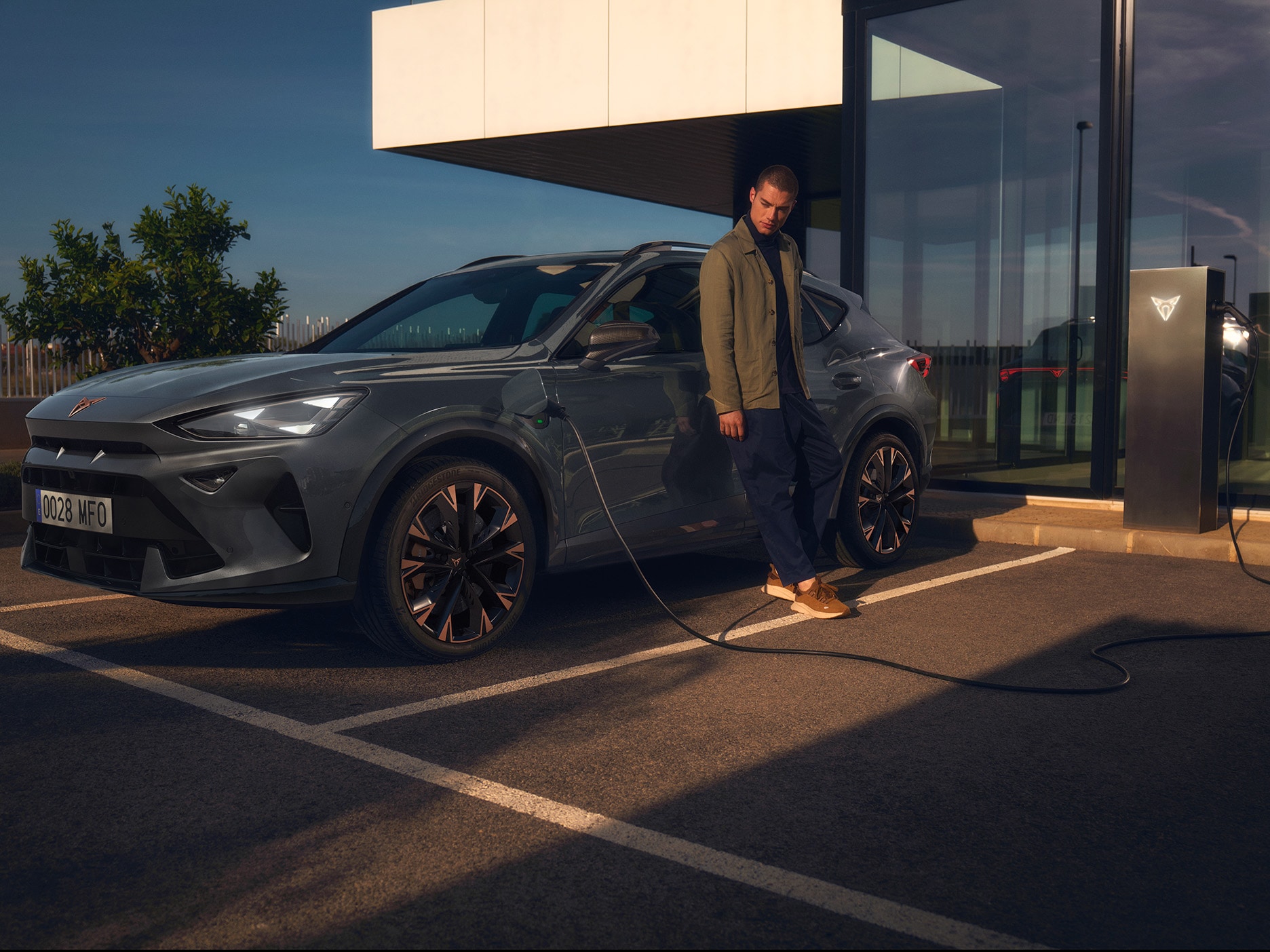 A man leaning against a CUPRA Formentor while it is parked and plugged in to a charging point.