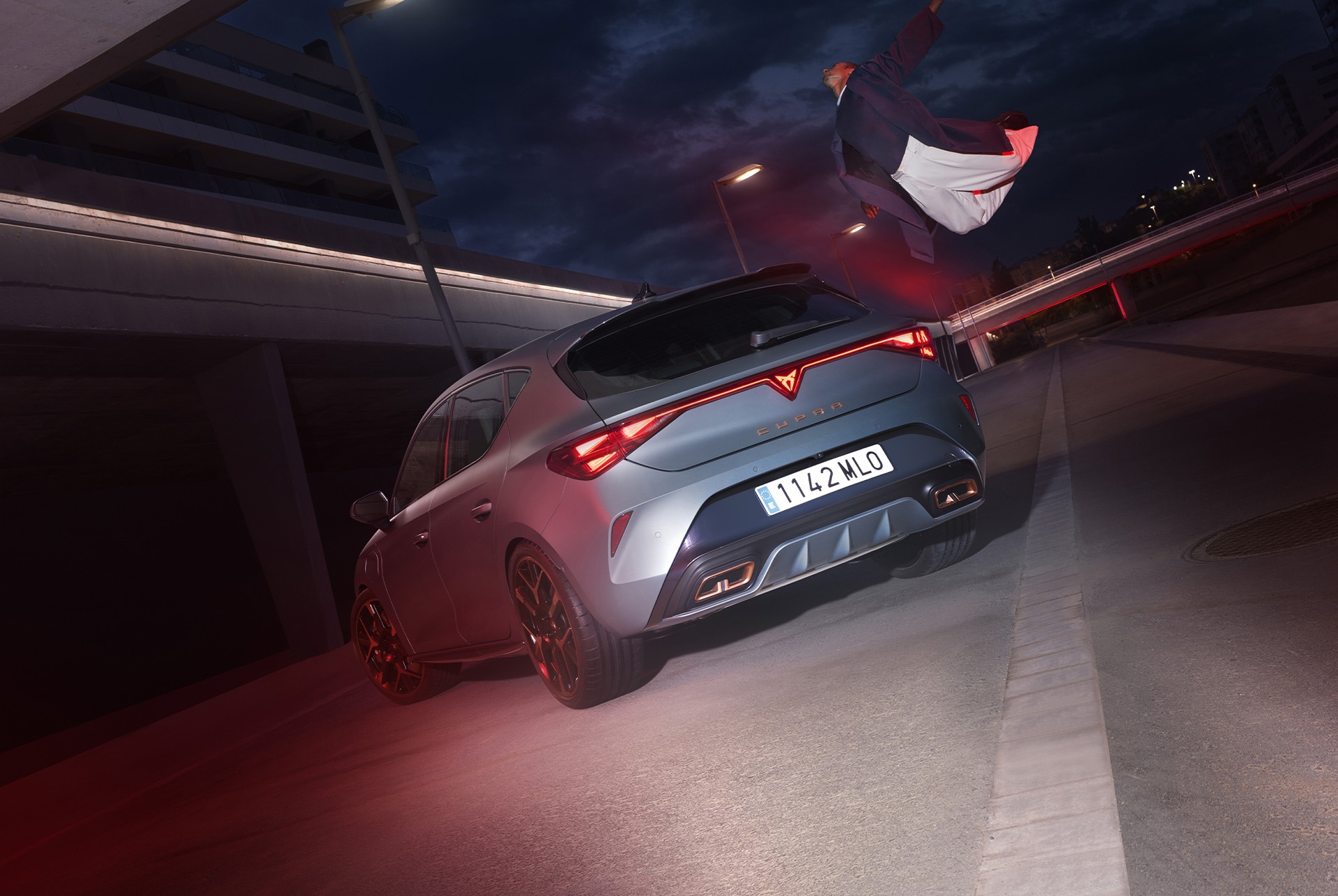 Hero angle, 3/4 rear view of the CUPRA Leon in Enceladus Grey Matt. The vehicle is parked in a urban setting at night, and there's a person next to it.