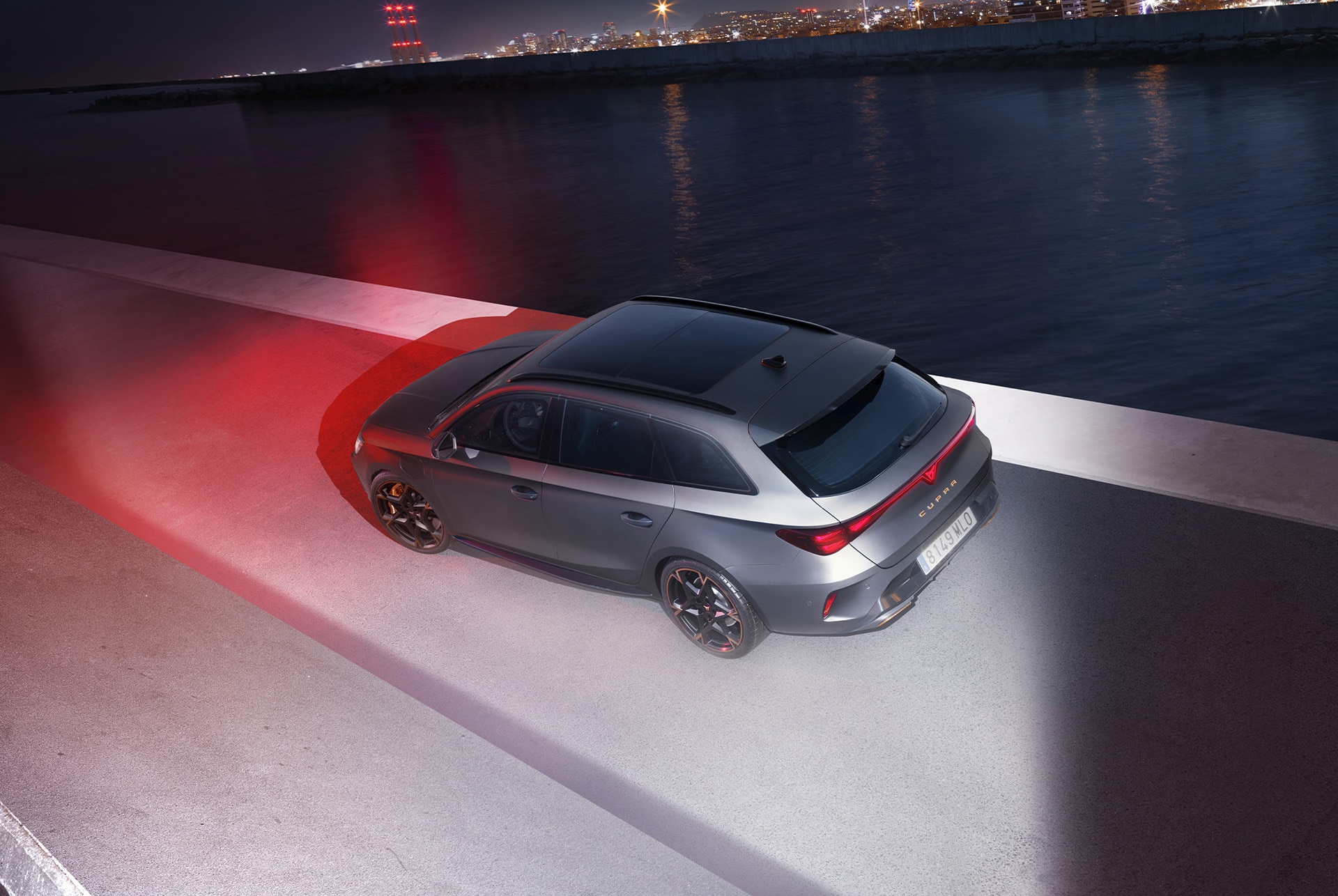 3/4 rear view of the CUPRA Leon Estate in Magnetic Tech Matt Grey with 19-inch alloy wheels in Polar Copper, at night, in a urban seaside landscape.