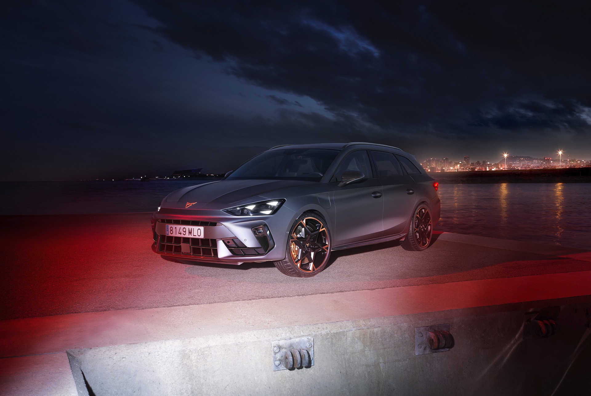 3/4 front view of the CUPRA Leon Estate in Magnetic Tech Matt Grey with an urban seaside backdrop at night, showcasing 19-inch alloy wheels in Polar Copper.