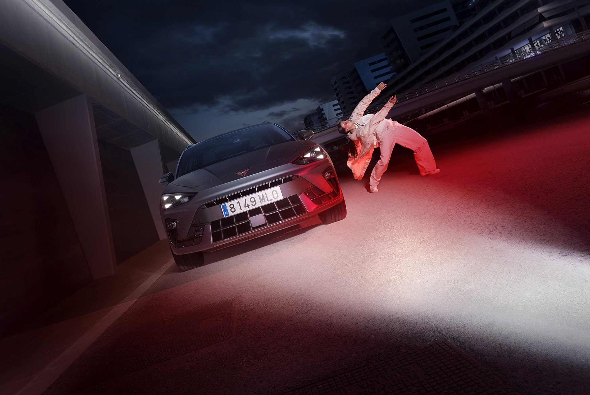 Front view of the CUPRA Leon Estate in Magnetic Tech Matt Grey colour, riding at night in a city, and an individual next to the vehicle.