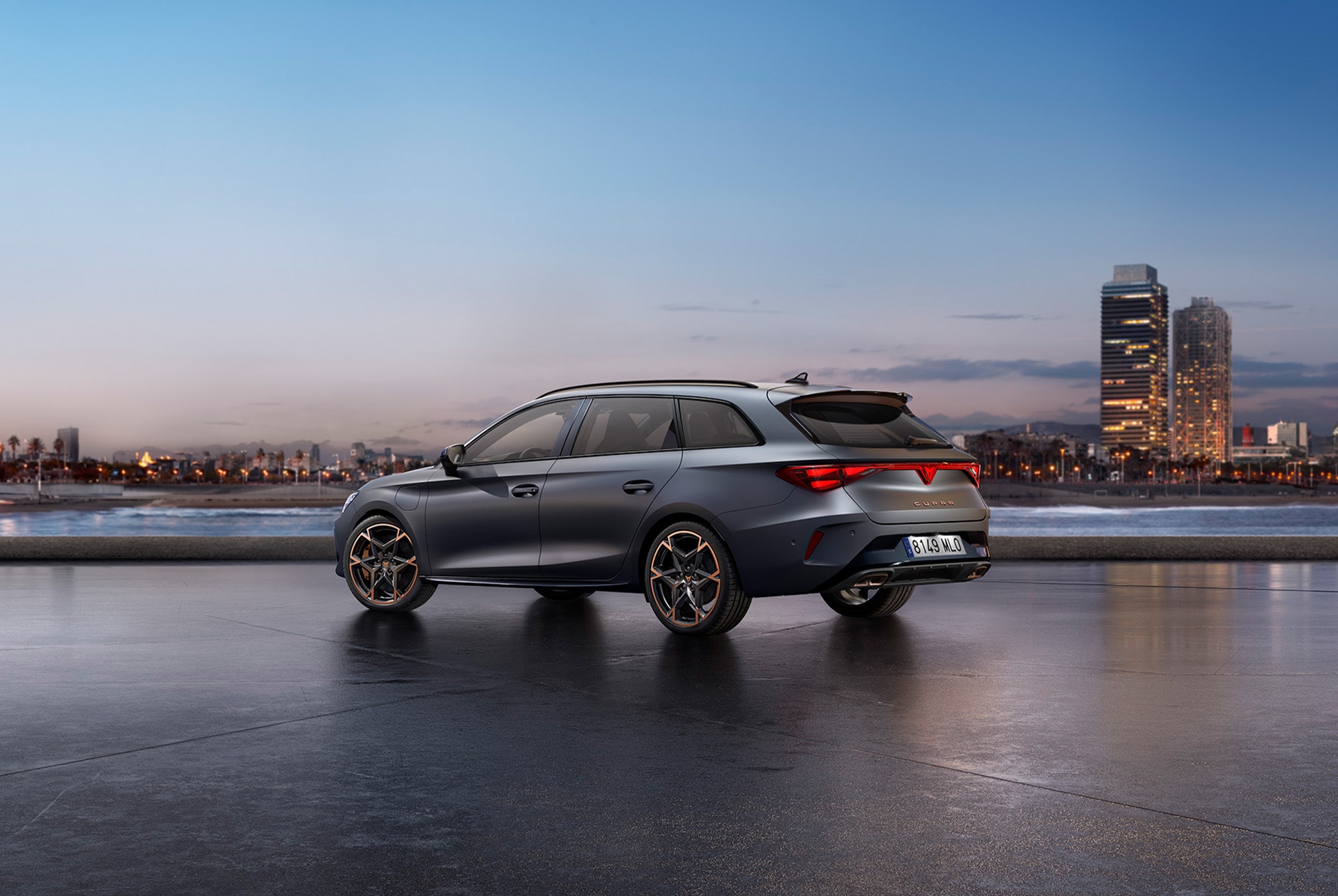 The sporty 2025 Cupra Leon Estate parked on concrete with a city skyline illuminated at twilight in the background. The vehicle is depicted in a matte grey finish.