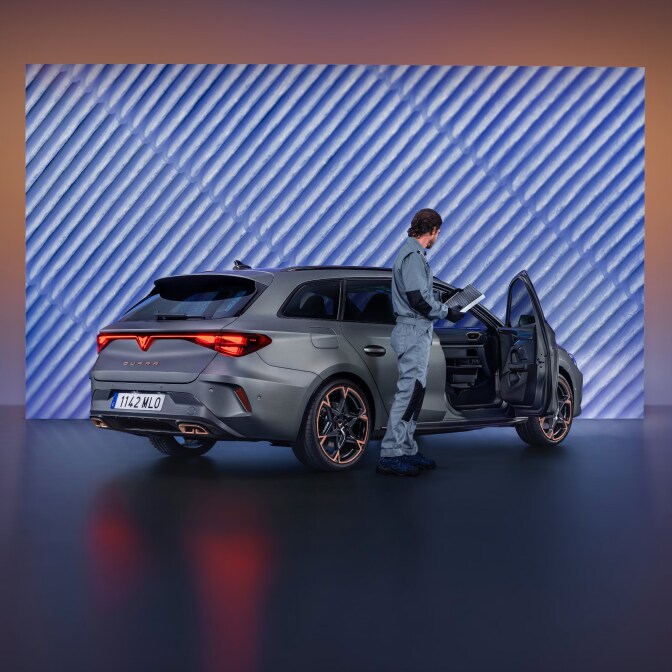 A technician in a grey jumpsuit inspecting the engine bay of a CUPRA Terramar in a studio; the background features a large screen displaying a close-up of metallic gears.