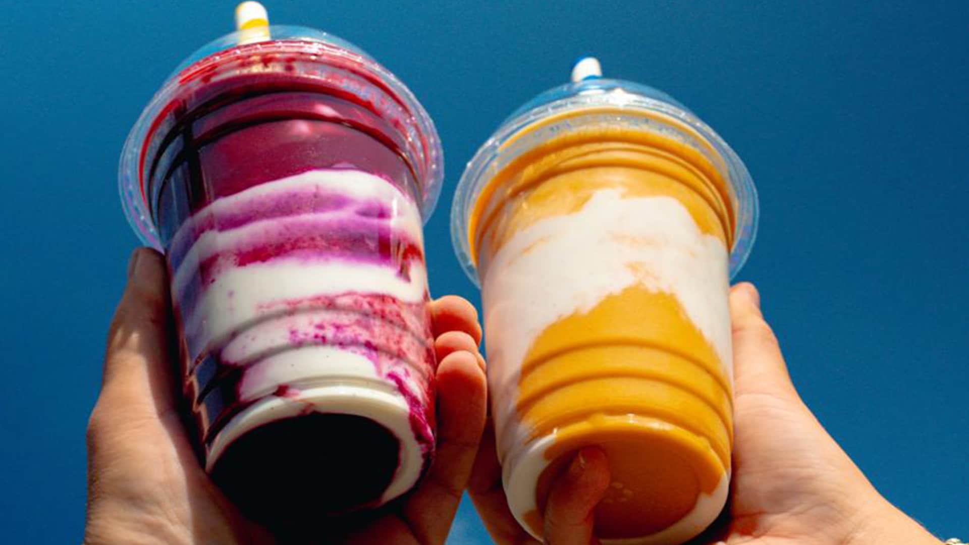 Two hands holding smoothies, one is purple and the other is yellow