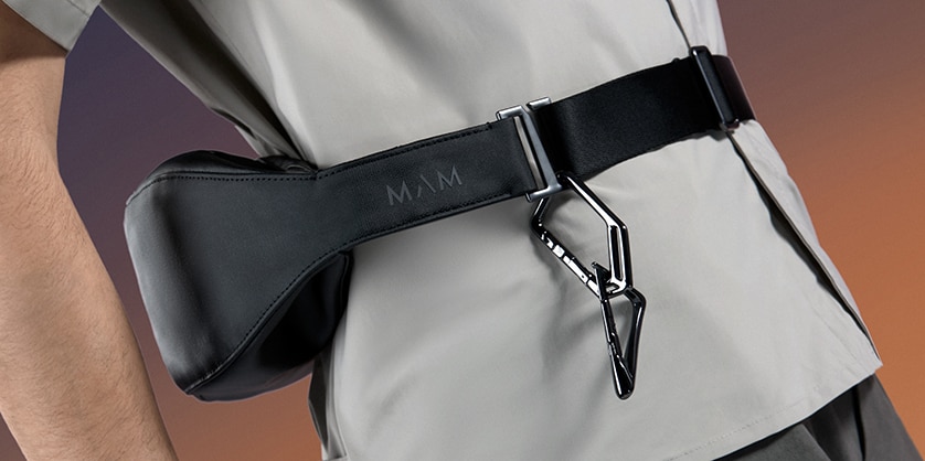 Black leather waist bag with adjustable belt and metal carabiner, worn around the hips over a grey outfit.