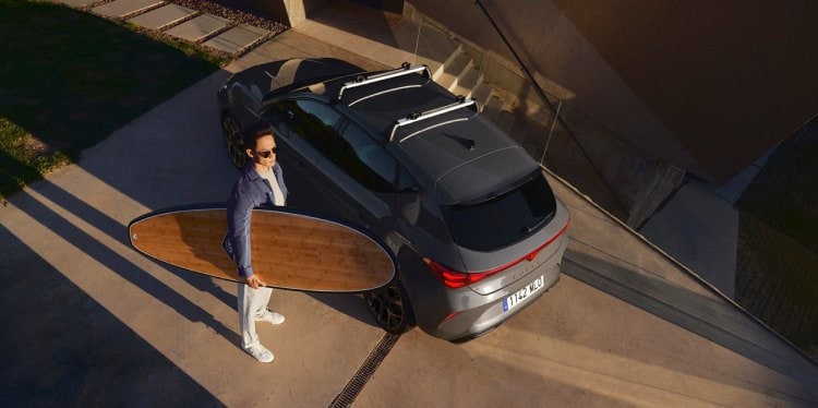 A person carrying a surfboard and standing next to a CUPRA Formentor