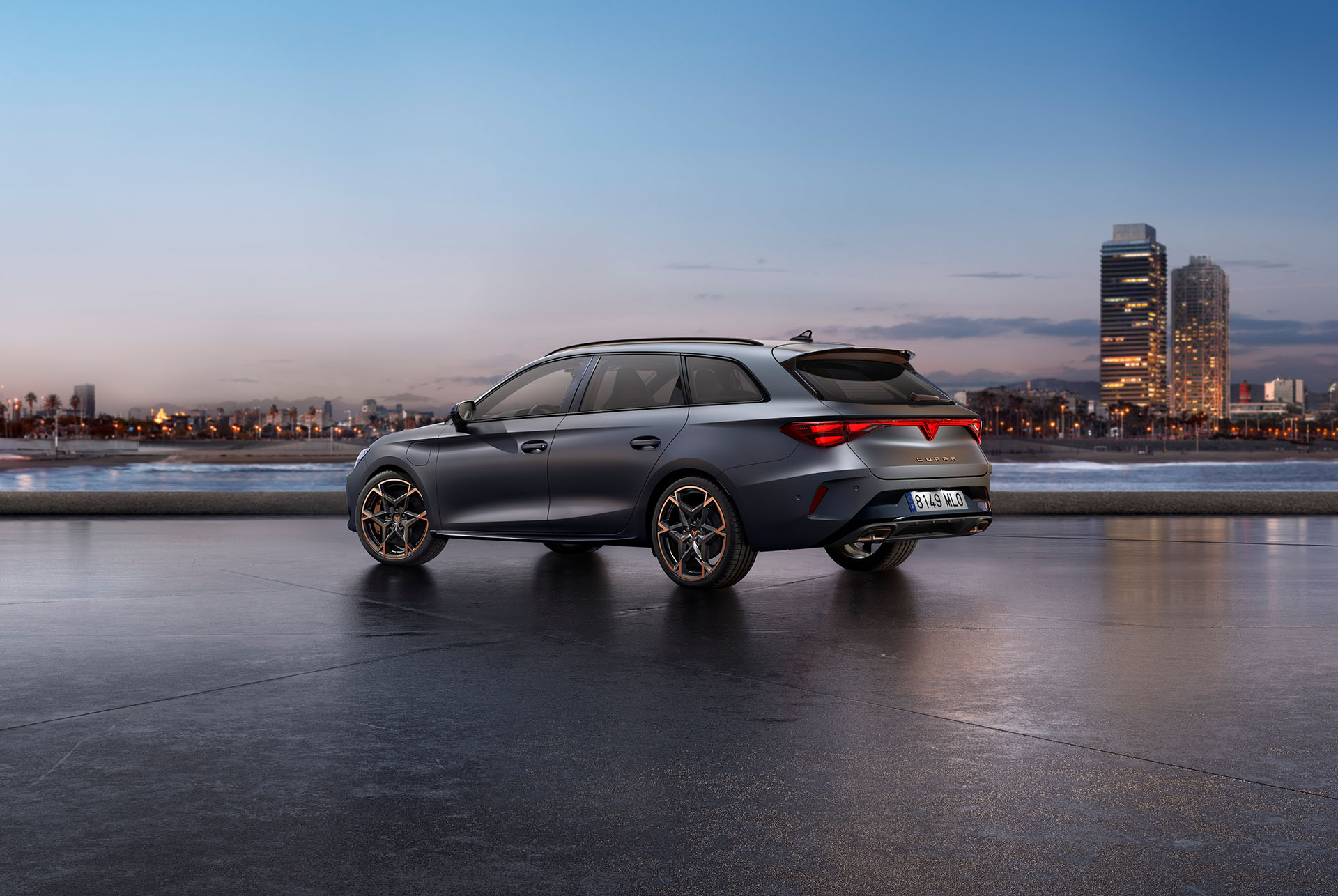 3/4 front view of the CUPRA Leon Estate in Magnetic Tech Matt Grey with an urban seaside backdrop at night, showcasing 19-inch alloy wheels in Polar Copper.