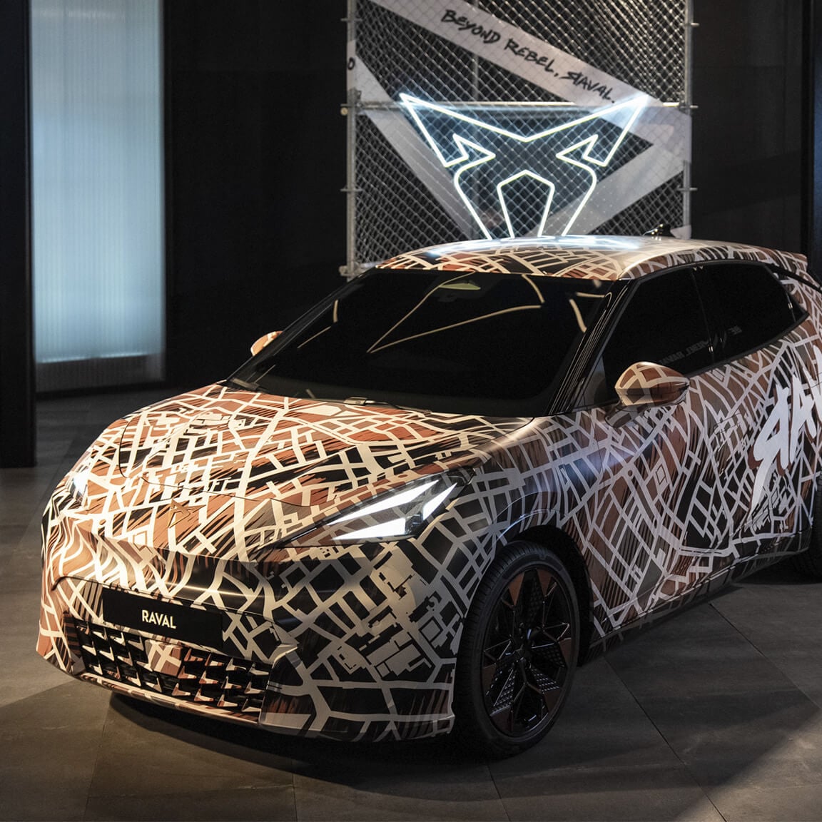 The CUPRA Raval, covered in a brown and white street map camouflage wrap, is displayed indoors. Behind the car is a chain-link fence panel with a glowing white CUPRA logo and the text "BEYOND REBEL, RAVAL.".