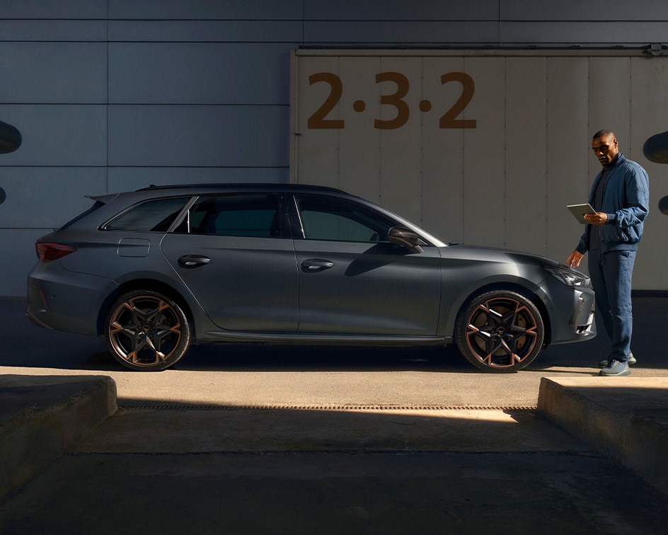 Two people standing between two parked CUPRA Terramar at twilight; a man on the right faces a woman on the left, who is standing near a dark green CUPRA Terramar and a wall-mounted charging station, while a purple Terramar is shown from the rear. 