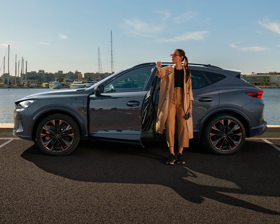 Two people standing between two parked CUPRA Terramar at twilight; a man on the right faces a woman on the left, who is standing near a dark green CUPRA Terramar and a wall-mounted charging station, while a purple Terramar is shown from the rear. 