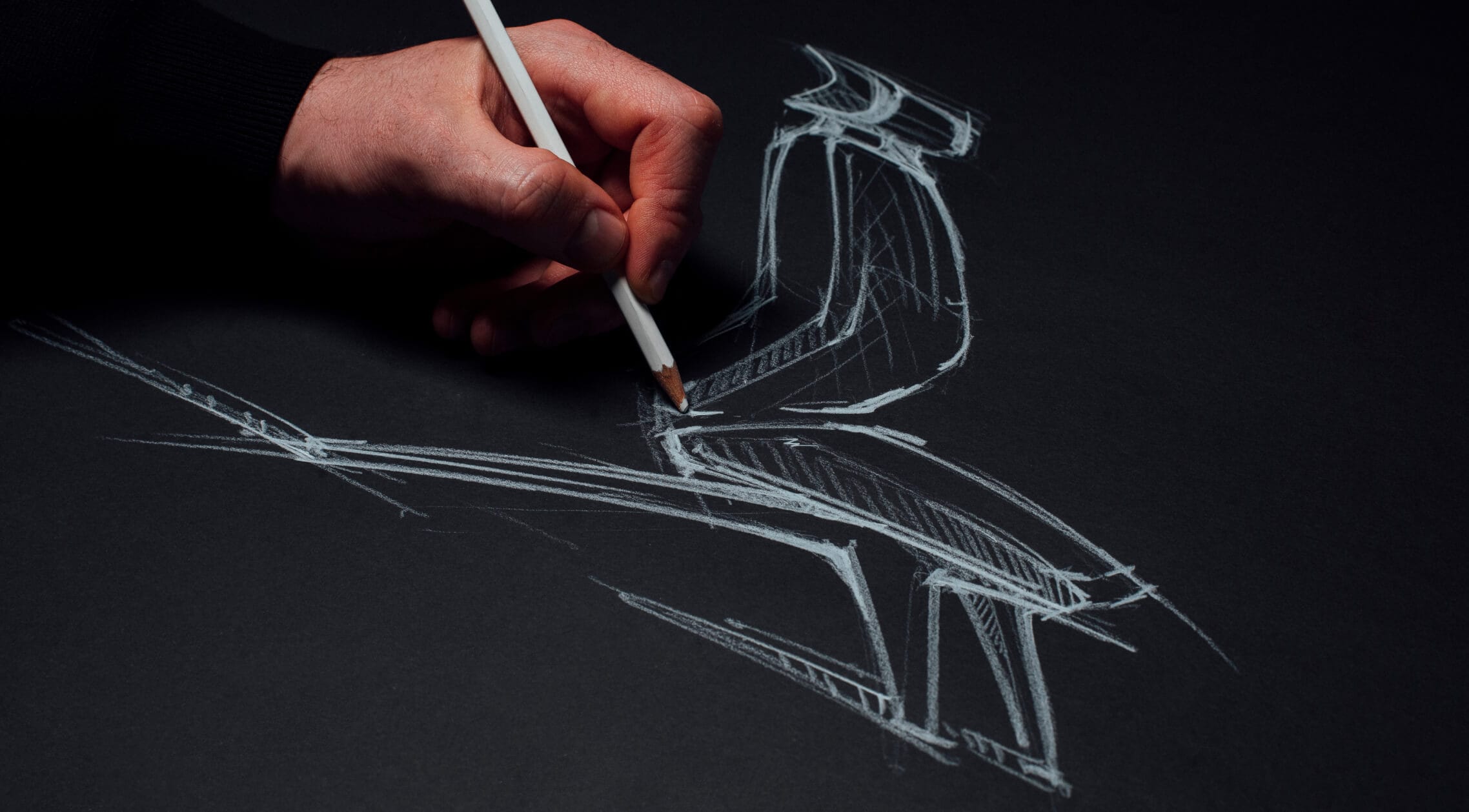 A hand holding a white pencil drawing a sketch of a chair with white lines on black paper. A hand holding a white pencil drawing a sketch of a chair with white lines on black paper.