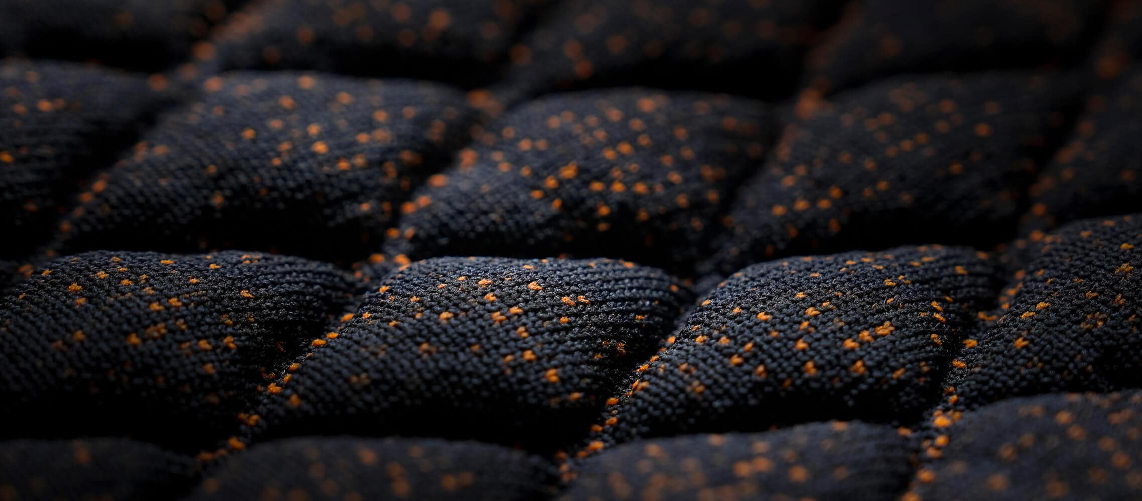 A close-up shot of a dark, quilted fabric with a raised diamond pattern and small orange flecks. A close-up shot of a dark, quilted fabric with a raised diamond pattern and small orange flecks.