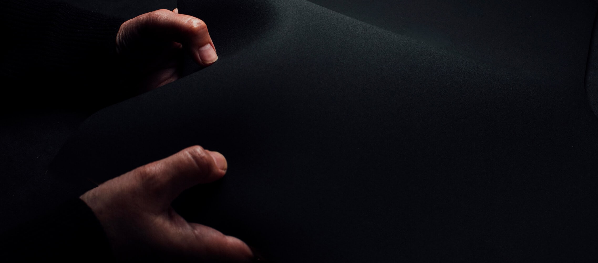 A close-up photograph of two hands holding a piece of black material with a dark background, blending with the material.