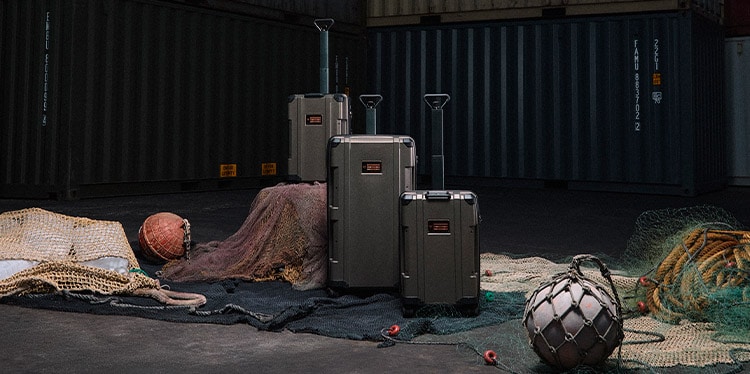 A set of three bronze-colored suitcases with black wheels and handles is the central focus of the image. The suitcases are arranged on top of what appears to be dark fabric and fishing nets. 